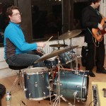 Man sits at drum set, other man stands playing guitar