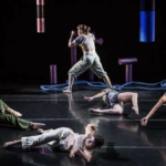 Four dancers on the ground in different poses, and one dancer standing up in the middle with their left leg bent ahead of them with their arms bent