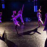 In image: five dancers hold their right arms up and their left legs outward on a stage.